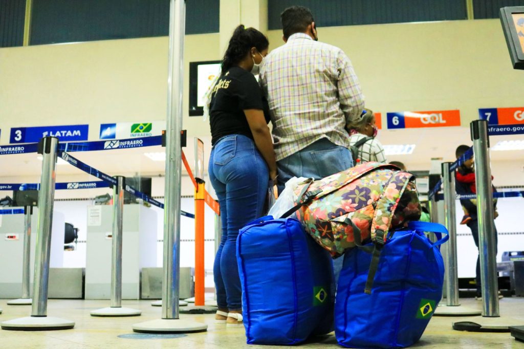 Casal está de costas na fila do check-in do aeroporto. Em destaque, na parte inferior, duas bolsas na cor azul e uma mochila
