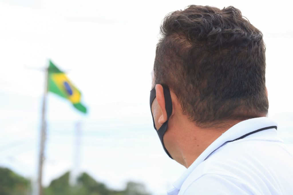 Homem está de costas para a imagem e observa bandeira do Brasil, ao fundo, tremulando